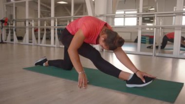 Pembe tişörtlü ve siyah taytlı genç bir kız spor salonundaki paspasın üzerine uzanıyor ve bacağını yarım sicimle eğiyor. Yavaş çekim