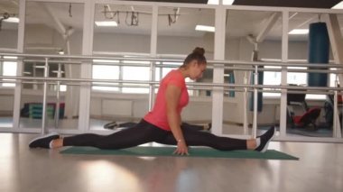 Çekici bir genç kız modern bir spor salonundaki bir paspasın üzerindeki boylamsal bir sicimin üzerine oturur. Aynanın arka planına karşı. Yavaş çekim