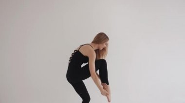 Giyim kuşamında güzel bir genç kız izole edilmiş beyaz bir stüdyoda esneyerek bacağını kaldırmaya çalışıyor. Yan görüş. Yavaş çekim