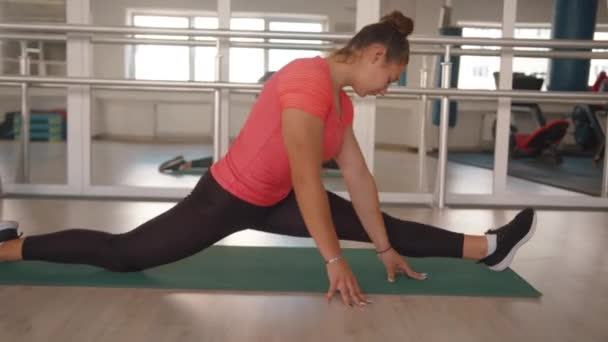 Симпатична молода дівчина тренується в спортзалі і розтягує м'язи ніг, сидячи на поздовжньому розколі — стокове відео