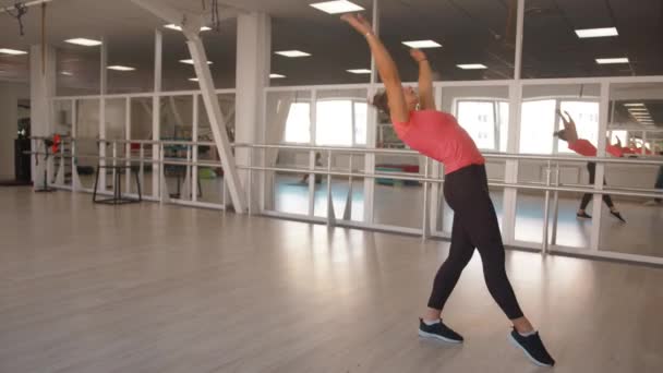 Piękna młoda dziewczyna w różowej koszulce i czarnych legginsach robi salto na prostych ramionach przed lustrem w sali gimnastycznej — Wideo stockowe