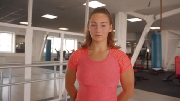 Een aangenaam jong meisje staat in de sportschool tegen de achtergrond van het trainen van bezoekers en straalt haar haar. Langzame beweging — Stockvideo