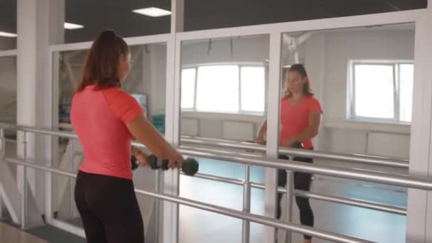 Ένα νεαρό κορίτσι που εκπαιδεύεται στο γυμναστήριο και εκτελεί αυξήσεις βραχίονα με αλτήρες μπροστά από έναν καθρέφτη για να λειτουργήσει έξω τους δελτοειδείς μυς — Αρχείο Βίντεο