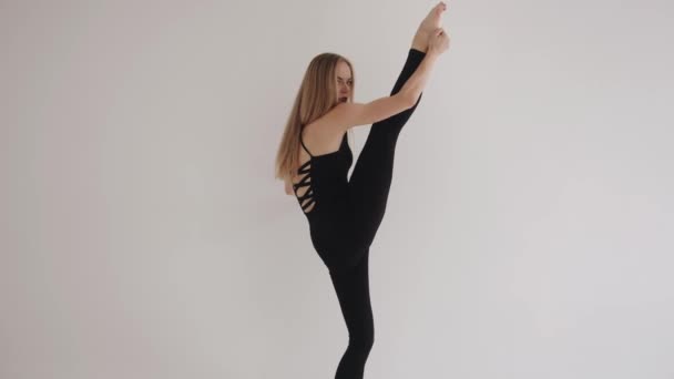 En ung flicka med ljus makeup i en vacker svart topp och leggings sträcker sig i en vit studio och höjer benet högt. Långsamma rörelser — Stockvideo