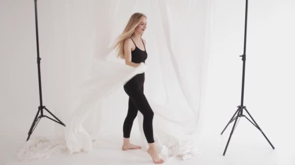 Spectacular young girl in black tight-fitting clothes with flowing hair stands wrapped in pieces of white tulle in the studio — Stock Video