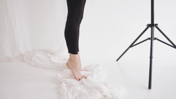 Una joven con polainas negras y pies descalzos haciendo estiramientos y se para de puntillas en una nube de tul blanco. Primer plano. Movimiento lento — Vídeos de Stock