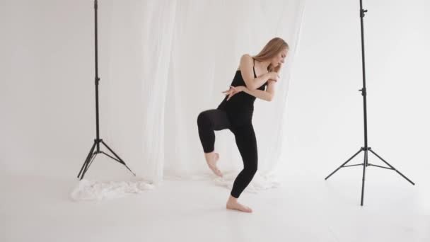 Uma jovem elegante está envolvida em alongamento e demonstra elementos de dança clássica contra um fundo de pedaços de tule branco — Vídeo de Stock