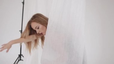Şehvetli genç kız, elleri beyaz tül parçalarının arka planında dans hareketleri yapıyor. Yavaş çekim