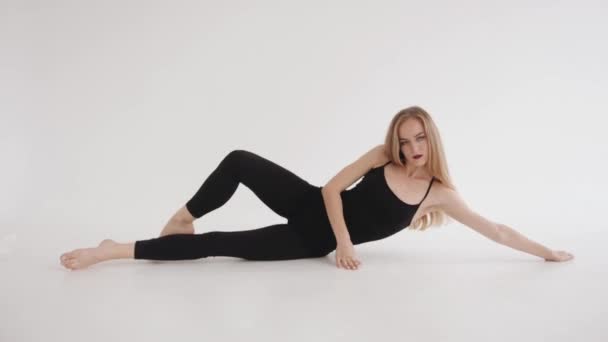 Een mooi jong meisje is bezig met stretchen in de studio en doet een longitudinale split liggend op de vloer. Langzame beweging — Stockvideo