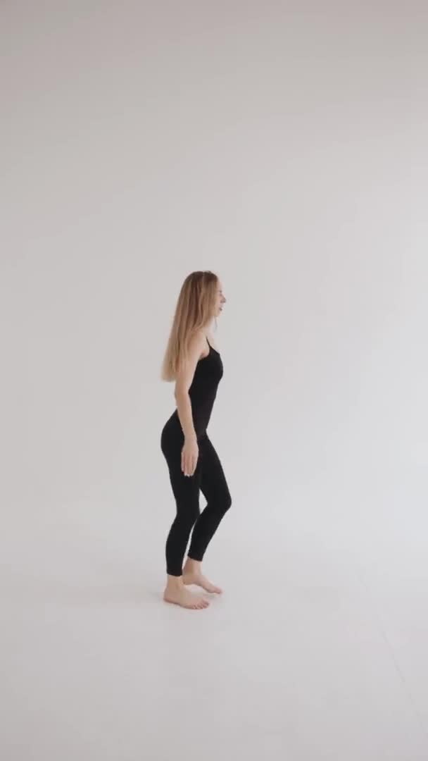 Charming young girl in a black top and leggings makes a ballet attitude jump in the studio on a white background. Vertical video — Stock Video
