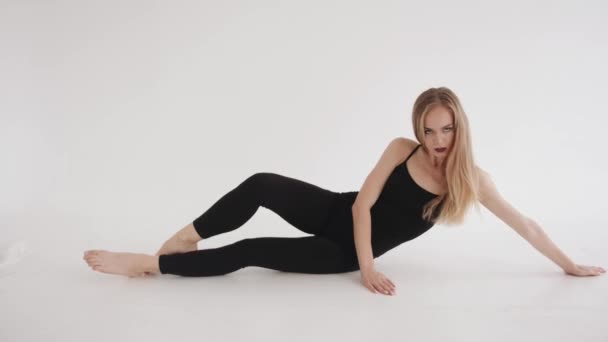 Sinnliches junges blondes Mädchen in schwarzer enger Sportbekleidung, das in einem weißen Studio auf dem Boden liegend Beinstreckung macht. Zeitlupe — Stockvideo
