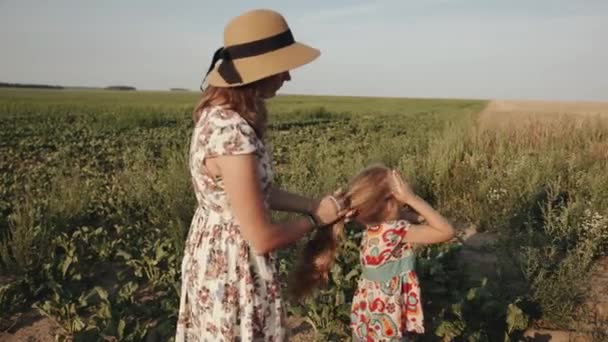 En mamma i sommarklänning och en hatt gör sina döttrar hår genom att binda en hästsvans på huvudet medan hon vilar på landet — Stockvideo