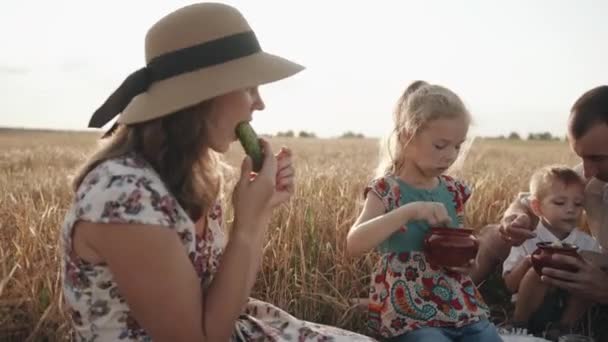 Jeune famille heureuse avec des enfants se reposant sur un pique-nique dans un champ de blé et mangeant de délicieux aliments. Concept d'amour et de soins — Video