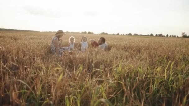 Pais com sua filha e filho em um piquenique em um campo de trigo com um cesto de alimentos e bebidas. Feliz conceito de família. Movimento lento — Vídeo de Stock