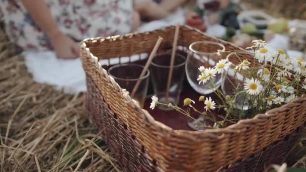 Cesto di vimini con bicchieri di vino e succo di frutta e con un mazzo di margherite su sfondo sfocato di un picnic in famiglia in un campo di grano. Rallentatore — Video Stock