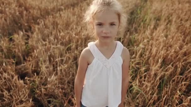 Portrait d'une jolie petite fille blonde aux cheveux longs en tunique blanche dans un champ de blé. Mouvement lent — Video