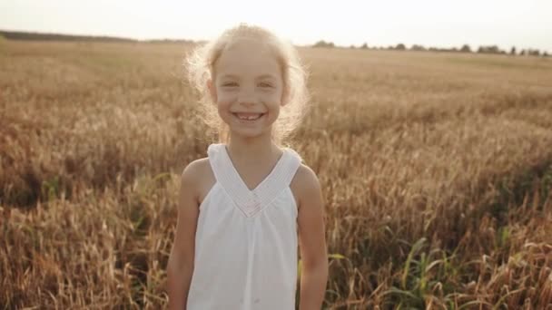 Seorang gadis TK ceria dengan gigi depan yang hilang berdiri di ladang gandum dan wajan di depan kamera. Gerakan lambat — Stok Video
