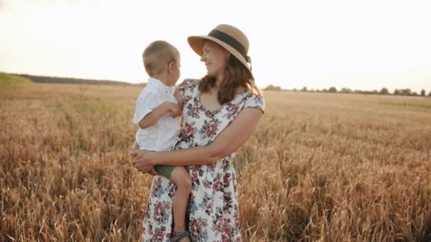 Moeder in een zomerjurk en een strohoed houdt haar zoontje in haar armen in een tarweveld en plaagt hem. Langzame beweging — Stockvideo