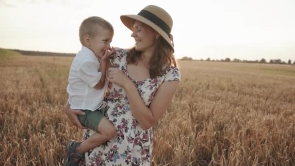 O mamă într-o rochie de vară și într-o pălărie de paie își ține fiul în brațe într-un câmp de grâu și îl gâdilă. Slow motion — Videoclip de stoc