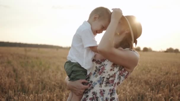 Una madre in cappello sta in un campo di grano con il suo piccolo figlio tra le braccia sullo sfondo del sole bacia e lo solletica. Rallentatore — Video Stock