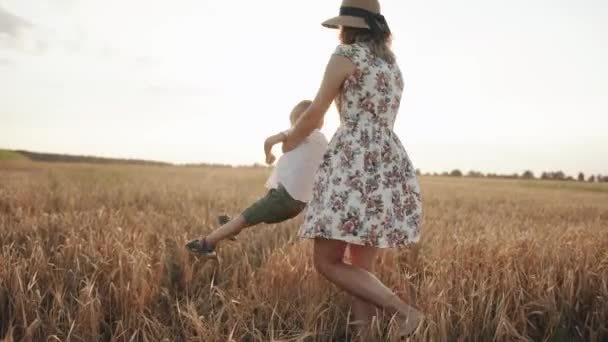 Seorang wanita muda yang cantik dengan gaun musim panas dan topi jerami berputar-putar dalam pelukan anak kecilnya di ladang gandum. Gerakan lambat — Stok Video