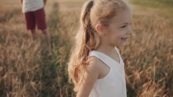 Een vrolijk klein meisje met ontbrekende voortanden die rotzooien en grimmig naar de camera kijken bij een familiepicknick in een tarweveld. Langzame beweging — Stockvideo