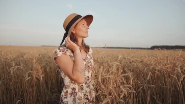 Uma jovem tenra está em um campo de trigo e olha para o céu colocando a mão sobre o chapéu. O conceito de liberdade e tranquilidade — Vídeo de Stock