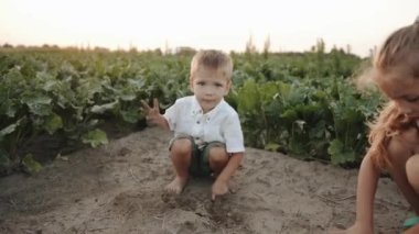 Küçük sevimli bir çocuk, yeşil taşıma aracının arasında sahada ablasıyla oynuyor ve elinde kumlar kazanıyor. Yavaş çekim