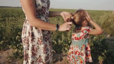 Yazlık elbiseli ve şapkalı bir anne kırsalda dinlenirken saçına at kuyruğu bağlayıp saçını yapıyor.