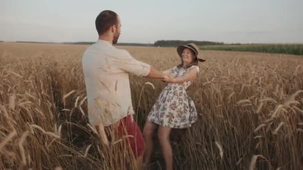 Şapkalı mutlu bir genç kadın ve kocası kırsalda gezmekten ve buğday tarlasının ortasında dönmekten zevk alıyorlar. Yavaş çekim — Stok video