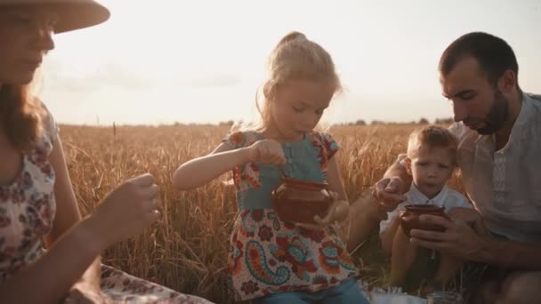 Μια νεαρή ευτυχισμένη οικογένεια με παιδιά χαλαρώνει σε ένα πικνίκ σε ένα χωράφι με σιτάρι και τρώει το φαγητό που έφεραν μαζί τους. Αργή κίνηση — Αρχείο Βίντεο
