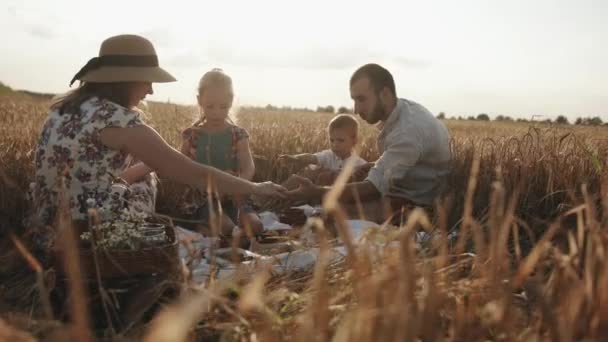 Přátelská rodina s dětmi sedí u improvizovaného stolu během pikniku na pšeničném poli za slunečného letního dne. Zpomal. Rodina se modlí společně — Stock video