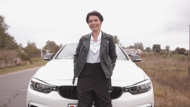 Siyah deri ceketli zarif bir genç kız üstü açık beyaz bir arabanın kaputuna yaslanıyor ve gülüyor. Yavaş çekim — Stok video