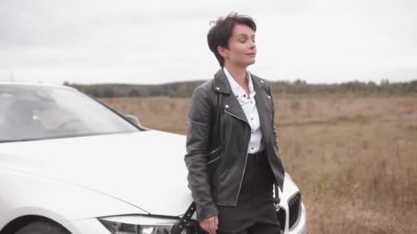 Charming young woman with short hair in a black leather jacket stands near a white car on a rural road. Slow motion — Stock Video