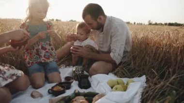 Çocuklu mutlu bir aile buğday tarlasında piknik yapıyor ve yanlarında getirdikleri yiyecekleri yiyor. Yavaş çekim