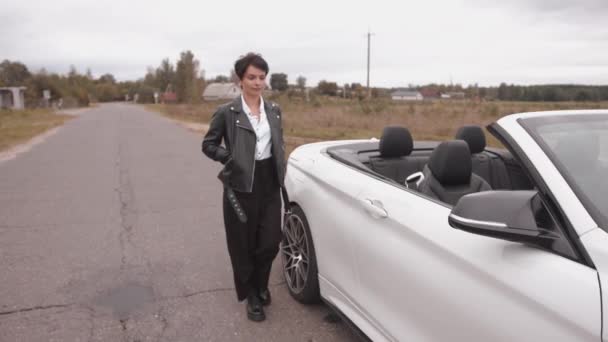 Uma jovem de luxo em uma jaqueta de couro preto vai ao longo de uma estrada rural e se aproxima de um conversível branco. Movimento lento — Vídeo de Stock