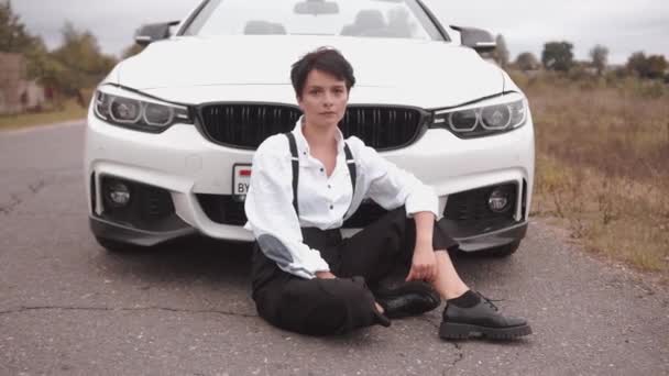 A striking young woman in a white shirt and black suspenders sits on the road in front of the hood of a white convertible. Slow motion — Stock Video