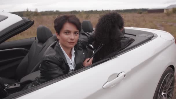 Mujer joven con estilo en una chaqueta de cuero negro se sienta en un convertible y acaricia un caniche grande. Movimiento lento — Vídeo de stock