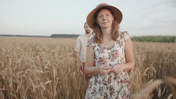 Un jeune homme à suivre à travers un champ de blé après sa femme dans une robe d'été et un chapeau sur une promenade en dehors de la ville. Mouvement lent — Video