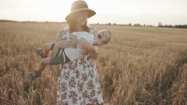 En vacker ung kvinna på ett vetefält håller sin son i sina armar och kittlar honom. Långsamma rörelser — Stockvideo