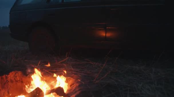 Une vieille voiture se tient sur un champ par une matinée nuageuse à côté d'un feu de joie dans une fosse et l'éblouissement du feu sur les portes. Concept de voyage et vacances — Video