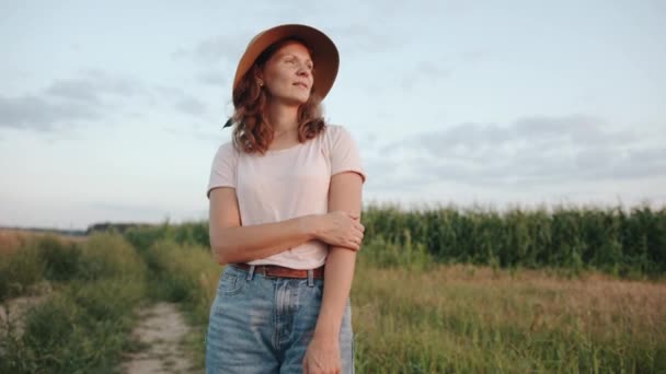 En trevlig ung kvinna står på vägen nära ett majsfält och justerar hatten på huvudet. Långsamma rörelser — Stockvideo