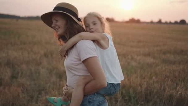 Şapkalı genç ve güzel bir kadın küçük kızını sırtında tutuyor ve buğday tarlasında onunla birlikte dönüyor. Yavaş çekim — Stok video