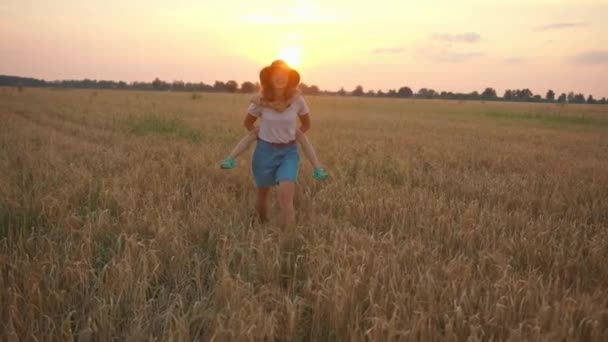 Une jeune femme souriante portant un chapeau et un short en jean porte sa fille sur le dos à travers un champ de blé au coucher du soleil. Mouvement lent — Video