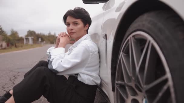 Una joven llamativa con una camisa blanca y tirantes negros se sienta en la carretera frente a la capucha de un convertible blanco. Movimiento lento — Vídeos de Stock