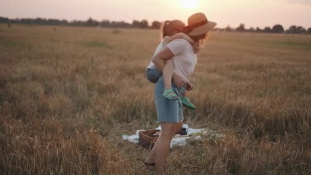 Un picnic familiar en un campo de trigo y una madre con su hija en la espalda girando y riendo. — Vídeos de Stock
