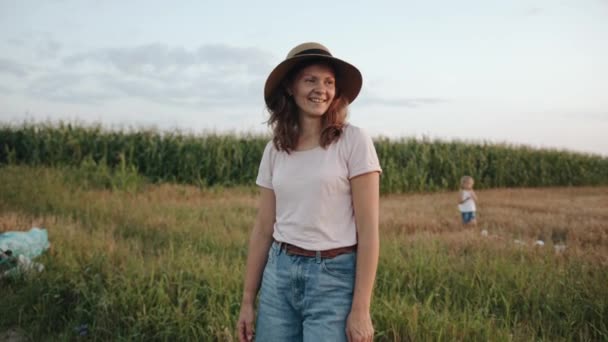 Μια ευχάριστη νεαρή γυναίκα στέκεται στο δρόμο κοντά σε ένα χωράφι με καλαμπόκια και προσαρμόζει το καπέλο της στο κεφάλι της. Αργή κίνηση — Αρχείο Βίντεο