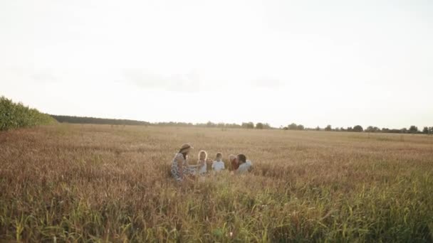Una madre con sombrero de paja y un padre en pantalones cortos rojos se sientan con sus hijos en un picnic familiar en un campo de trigo. Movimiento lento — Vídeo de stock