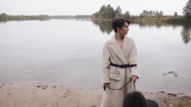 Kısa saçlı enfes bir genç kadın nehir kıyısında fino köpeğiyle yürüyor ve sıcak bir sonbahar gününün tadını çıkarıyor. Yavaş çekim