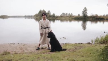 Beyaz önlüklü zarif genç bir kadın nehir kıyısında duruyor ve büyük siyah bir kanişi tasmasında tutuyor. Yavaş çekim